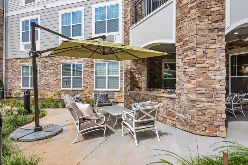 Outdoor patio area with cushioned chairs and a table under a large green umbrella, adjacent to a building with stone and siding exterior walls and multiple windows.