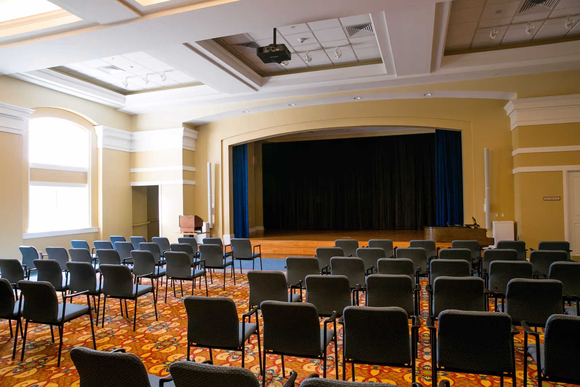 An empty auditorium or meeting room with rows of chairs facing a stage with a closed black curtain. The room has large windows letting in natural light, a colorful patterned carpet, and a ceiling with recessed lighting and a projector.