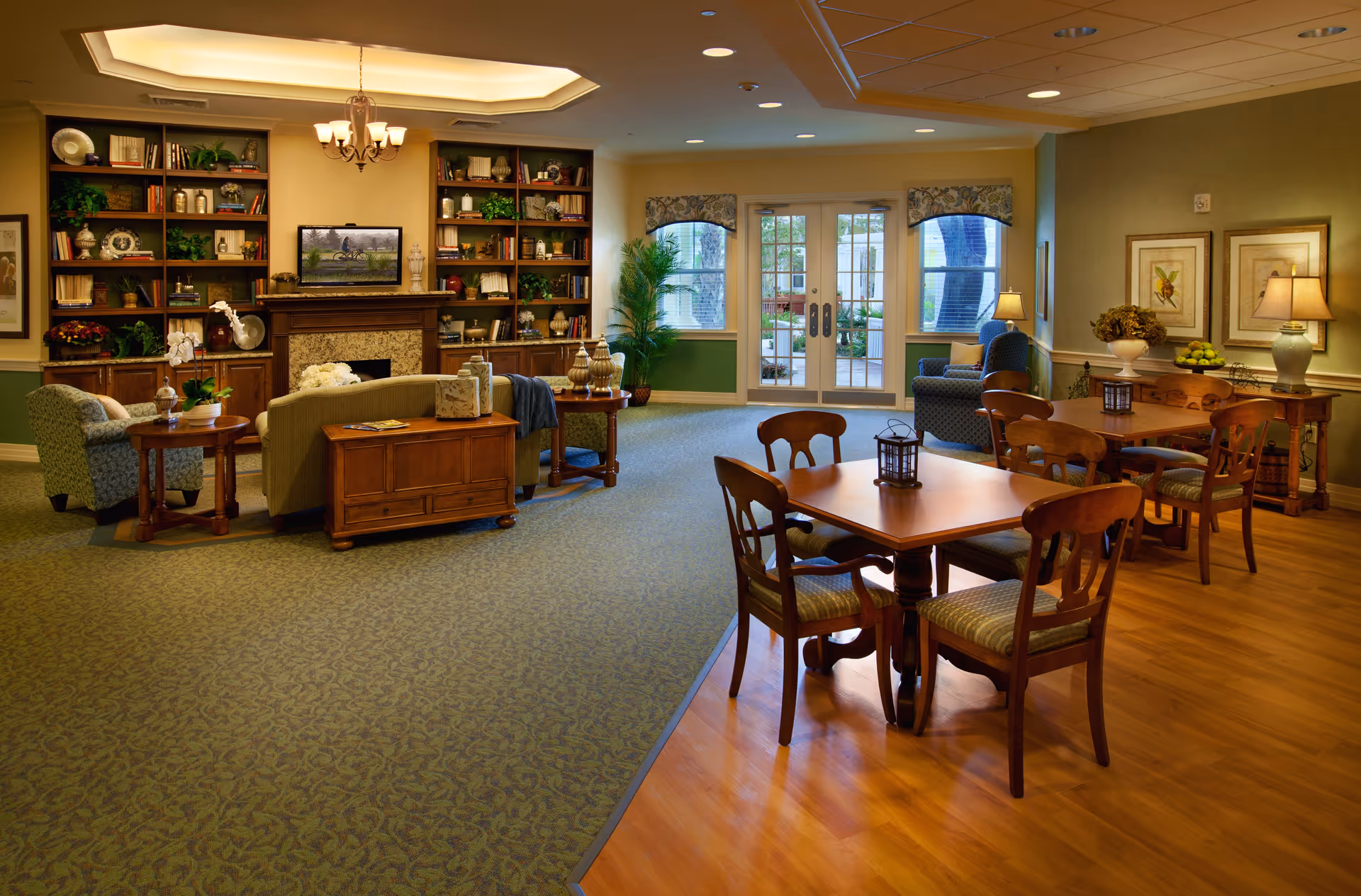 A cozy senior living facility common area with a seating arrangement including armchairs and a sofa around a fireplace with built-in bookshelves. There is a wooden dining table with chairs on a wood floor section, and large windows and glass doors letting in natural light. The room is warmly lit with lamps and a chandelier, decorated with plants, framed artwork, and decorative items.