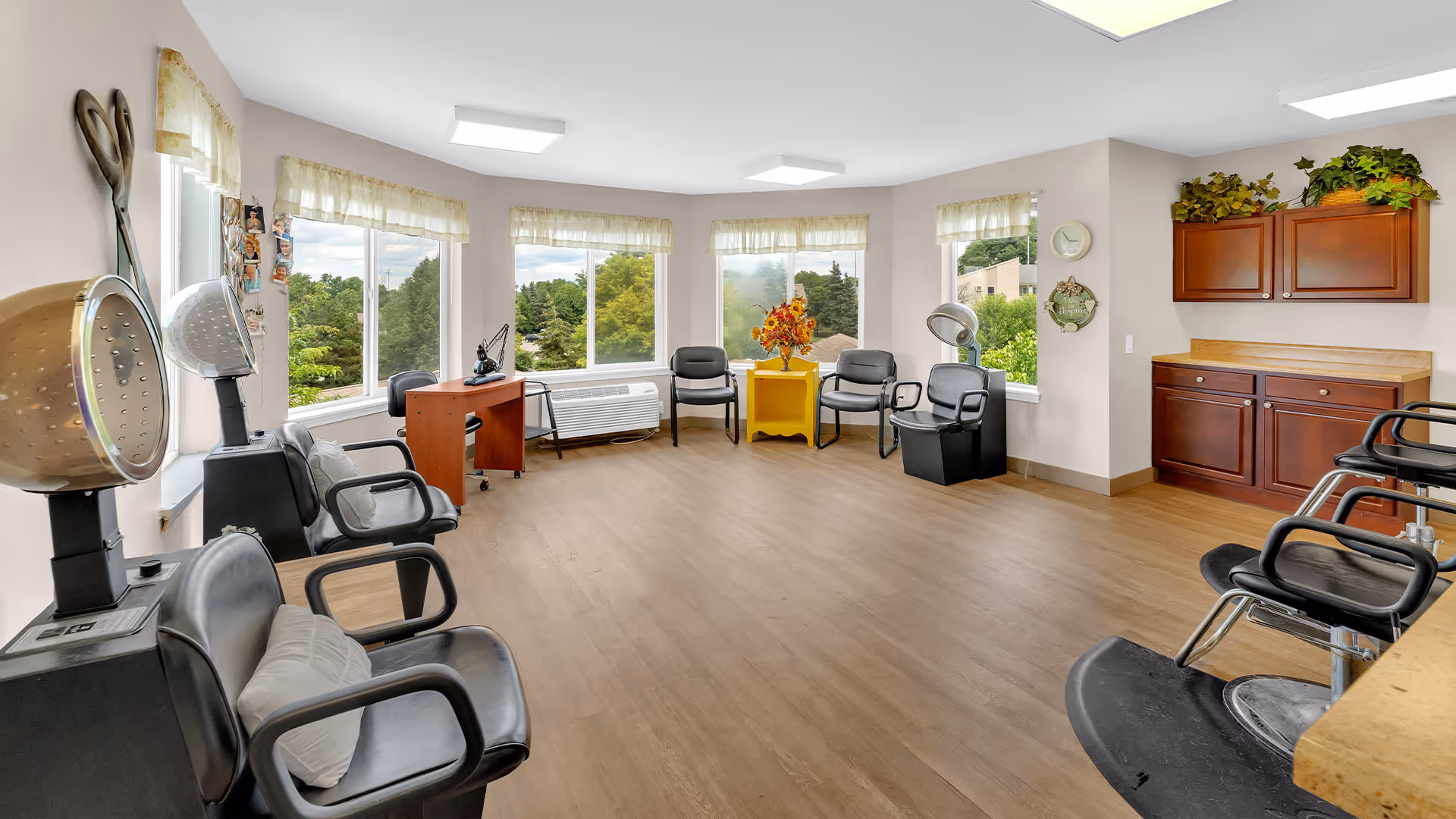 A bright and spacious salon area with multiple black salon chairs and hair drying stations. Large windows with sheer curtains let in natural light and offer a view of green trees outside. There is a small wooden desk with a telephone, a yellow side table with a flower arrangement, and wooden cabinets with plants on top along one wall.