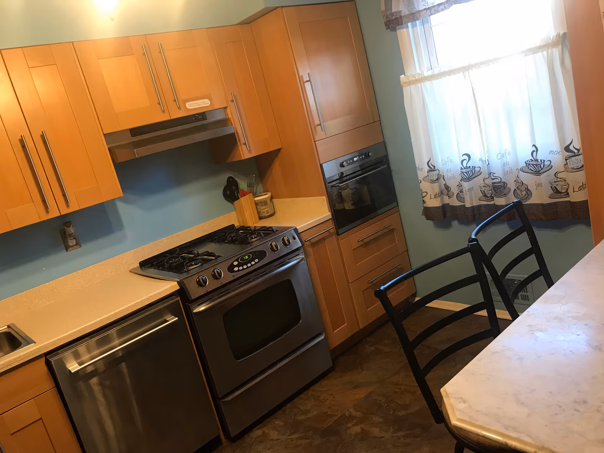 A kitchen with wooden cabinets, a stainless steel gas stove and oven, a dishwasher, and a built-in microwave. There is a window with a curtain featuring coffee cup designs, and two black chairs are positioned next to a marble countertop.