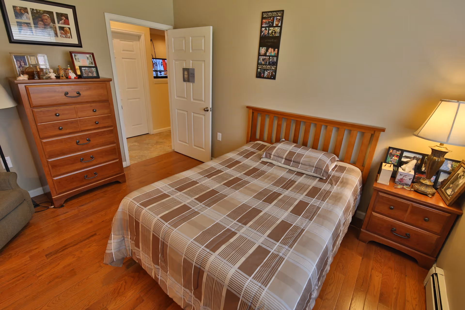 A bedroom with a wooden bed frame and a plaid bedspread in shades of brown and beige. There is a matching wooden nightstand with a lamp and framed photos on it to the right of the bed. To the left, there is a wooden dresser with more framed photos and decorative items on top. The room has hardwood floors and beige walls. An open door reveals a hallway with a television mounted on the wall.