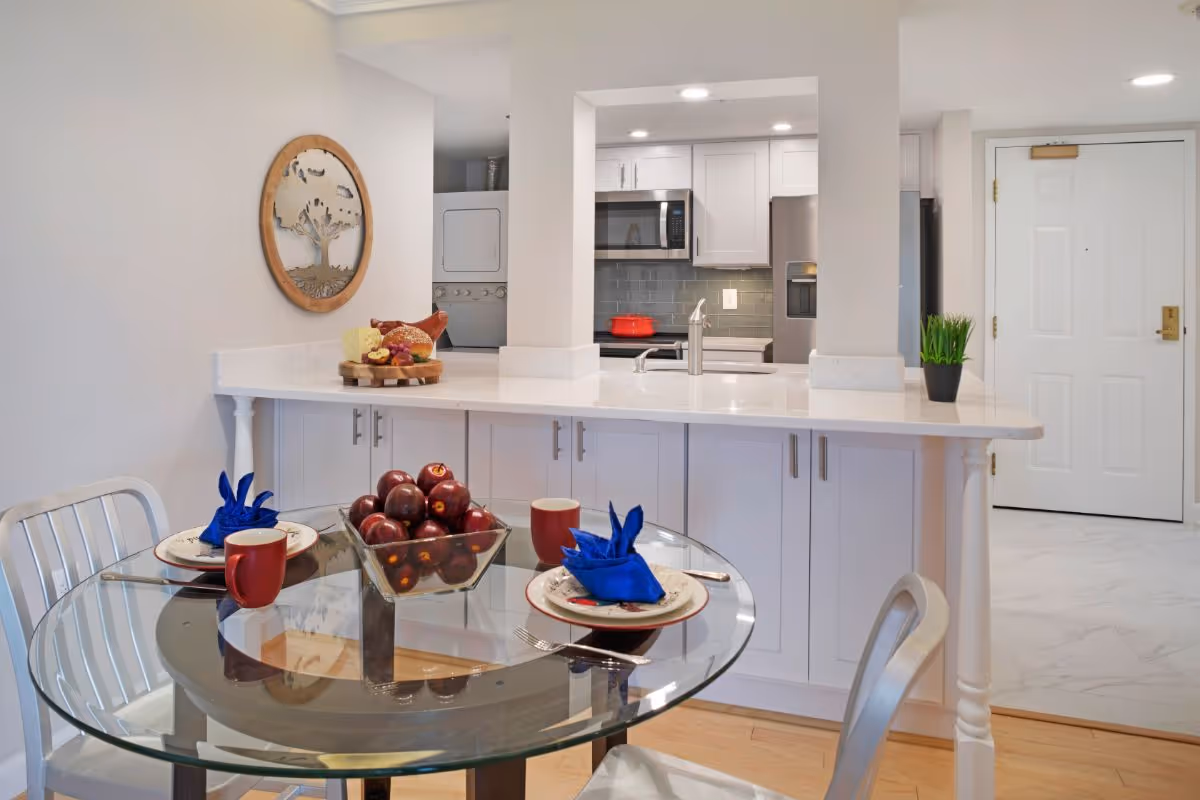 A modern kitchen and dining area with a glass-top round table set for two with red mugs and blue napkins. The kitchen features white cabinets, a stainless steel microwave and refrigerator, a red pot on the stove, and a small potted plant on the counter. A stacked washer and dryer are visible in a small alcove. The entry door is seen in the background.