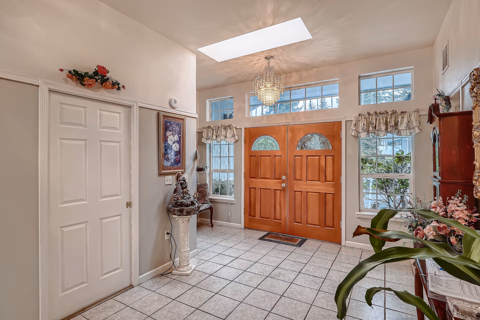 Bright tiled foyer with double wooden front doors, transom windows, a chandelier, and decorative plants and furniture.