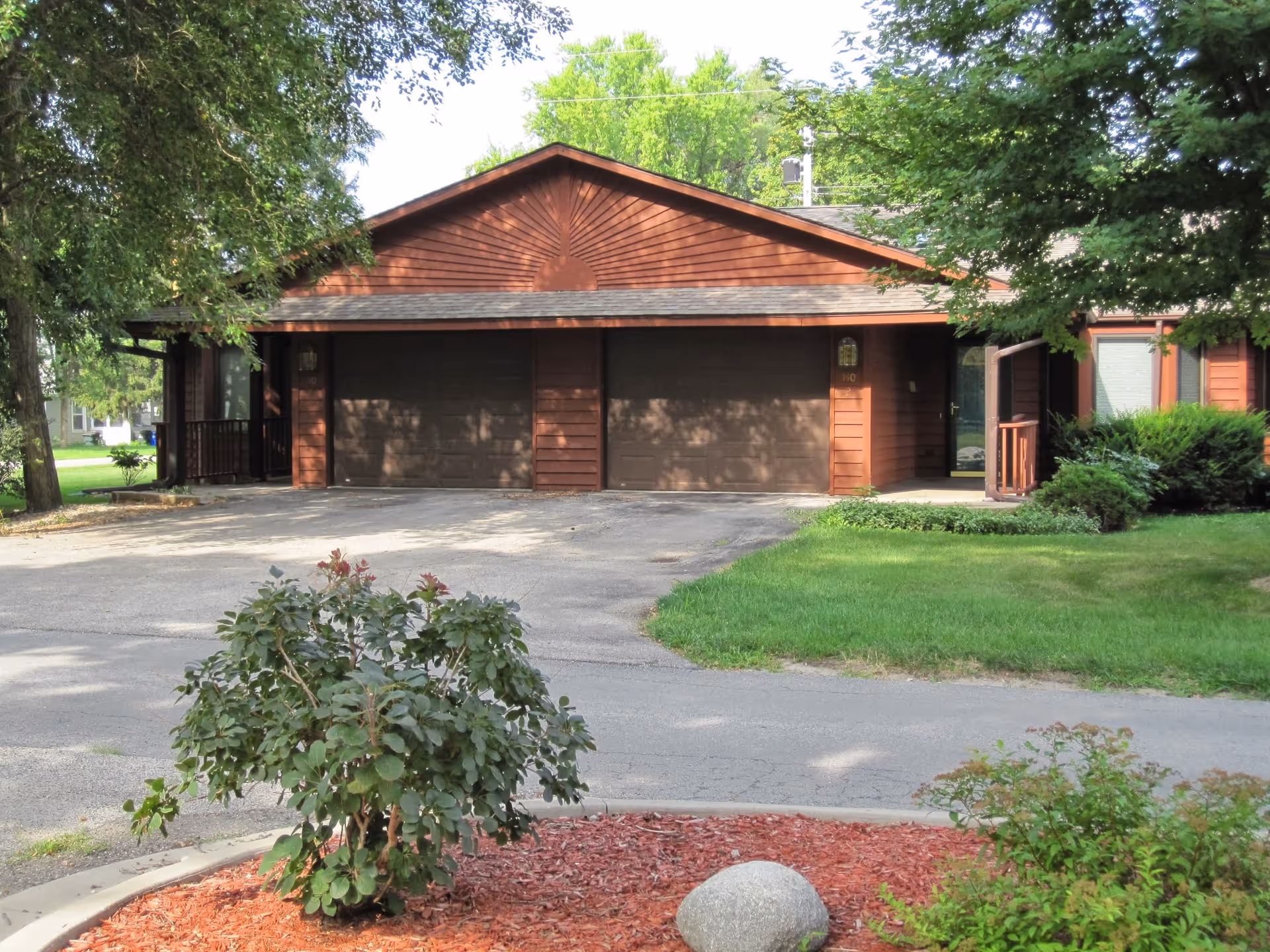 Exterior view of a single-story brown wooden building with two closed garage doors, surrounded by green trees and bushes, with a paved driveway and a landscaped area with mulch and a rock in front.