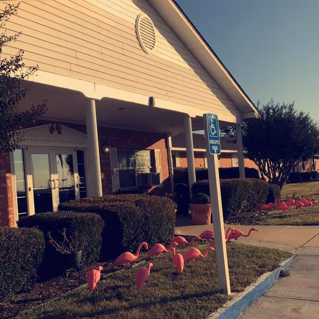 Exterior view of Colonial Park Manor showing the front entrance with a covered porch supported by white columns. There are neatly trimmed bushes and a row of pink plastic flamingos on the grass near the sidewalk. A handicapped parking sign is visible near the entrance.