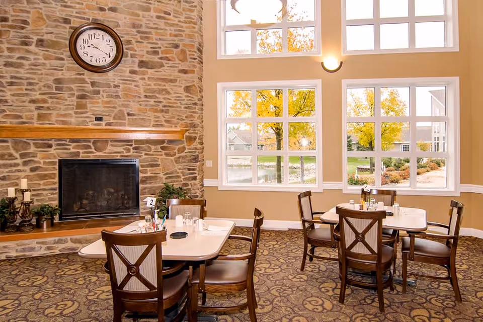 Dining room with tables and chairs, a large stone fireplace, and tall windows showing autumn trees outside.
