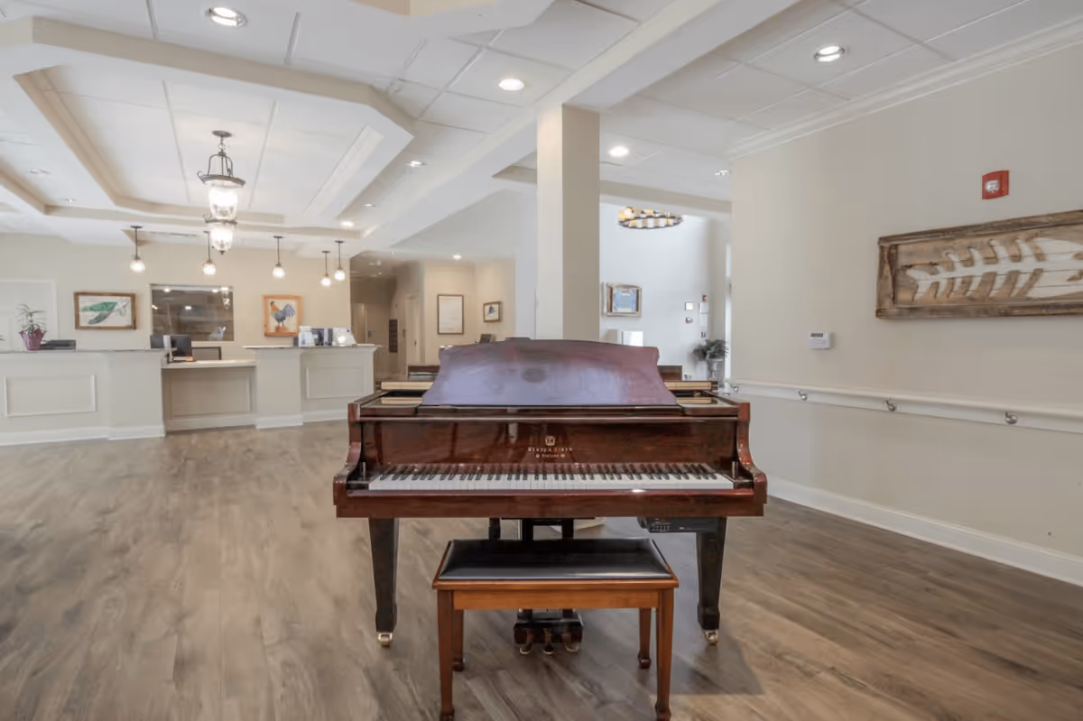 A bright senior living facility common area with a grand piano centered and a reception desk in the background.