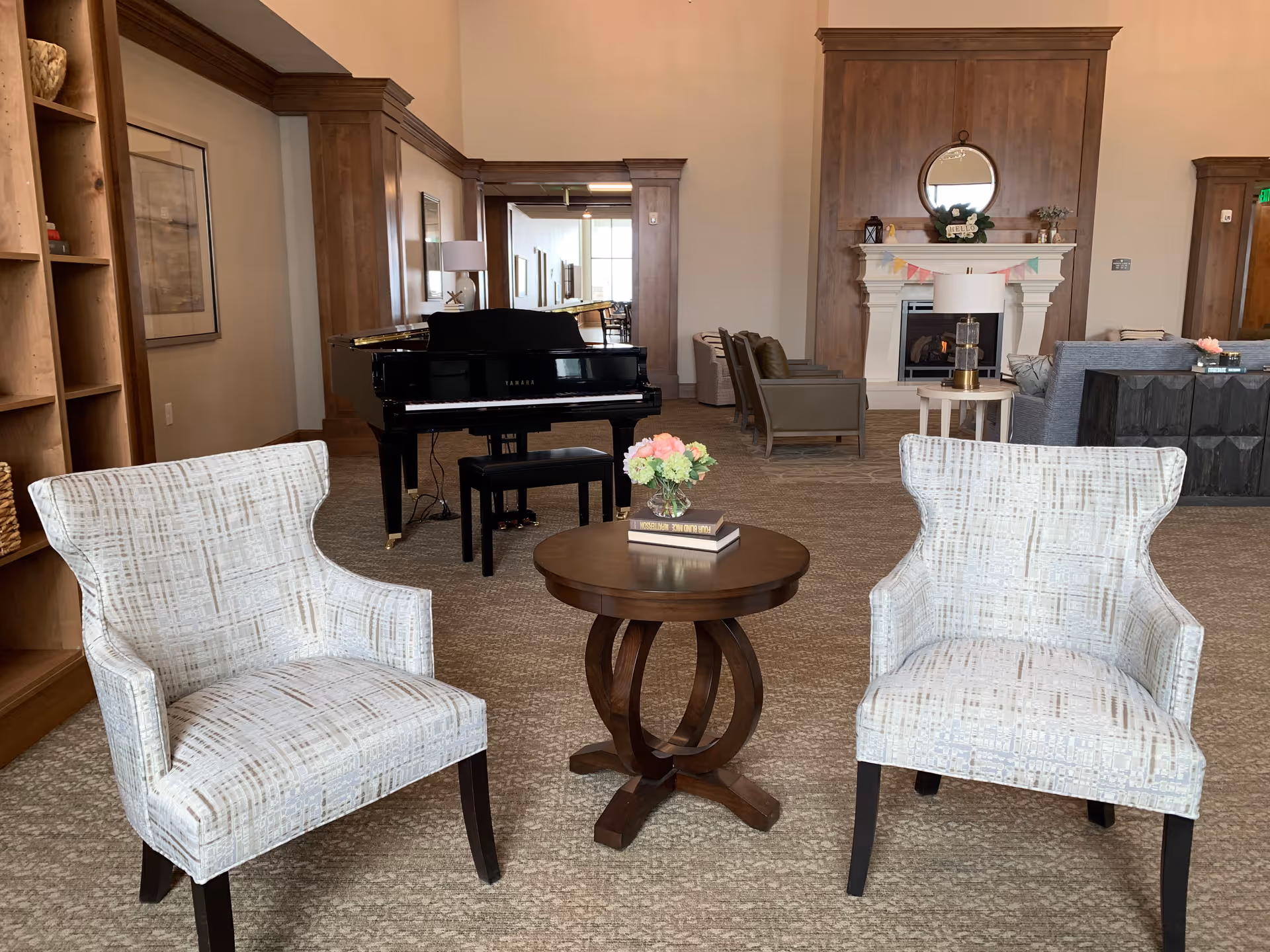 Two upholstered chairs flanking a round wooden table in a spacious common room with a grand piano and fireplace in the background.