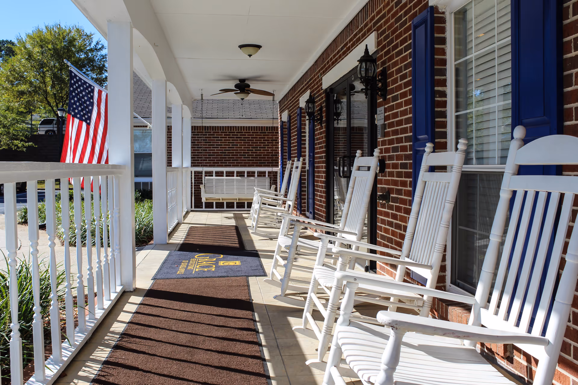 A sunny porch with white rocking chairs lined up against a brick wall with blue window shutters. The porch has a ceiling fan, hanging swing, and an American flag on the left side. There are brown mats on the tiled floor, one with the text 'Grace Senior Living'.