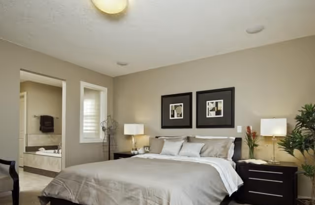 A neatly made bed with beige and white bedding in a bedroom. Two framed abstract artworks hang above the bed. There are two bedside tables with lamps and a plant on the right side. An open doorway reveals a bathroom with a bathtub and a towel hanging on the wall.