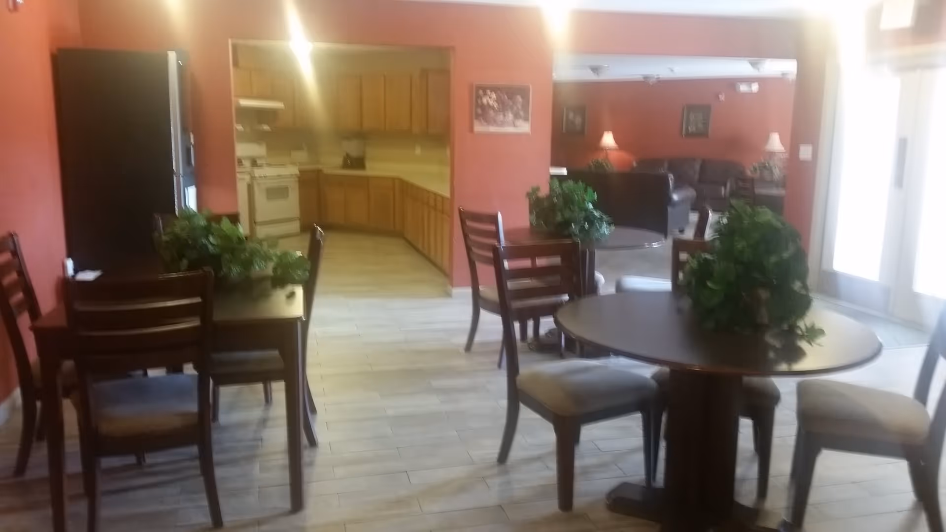 Community dining area with round and rectangular tables and chairs, potted plants, and a view into a kitchen and lounge.