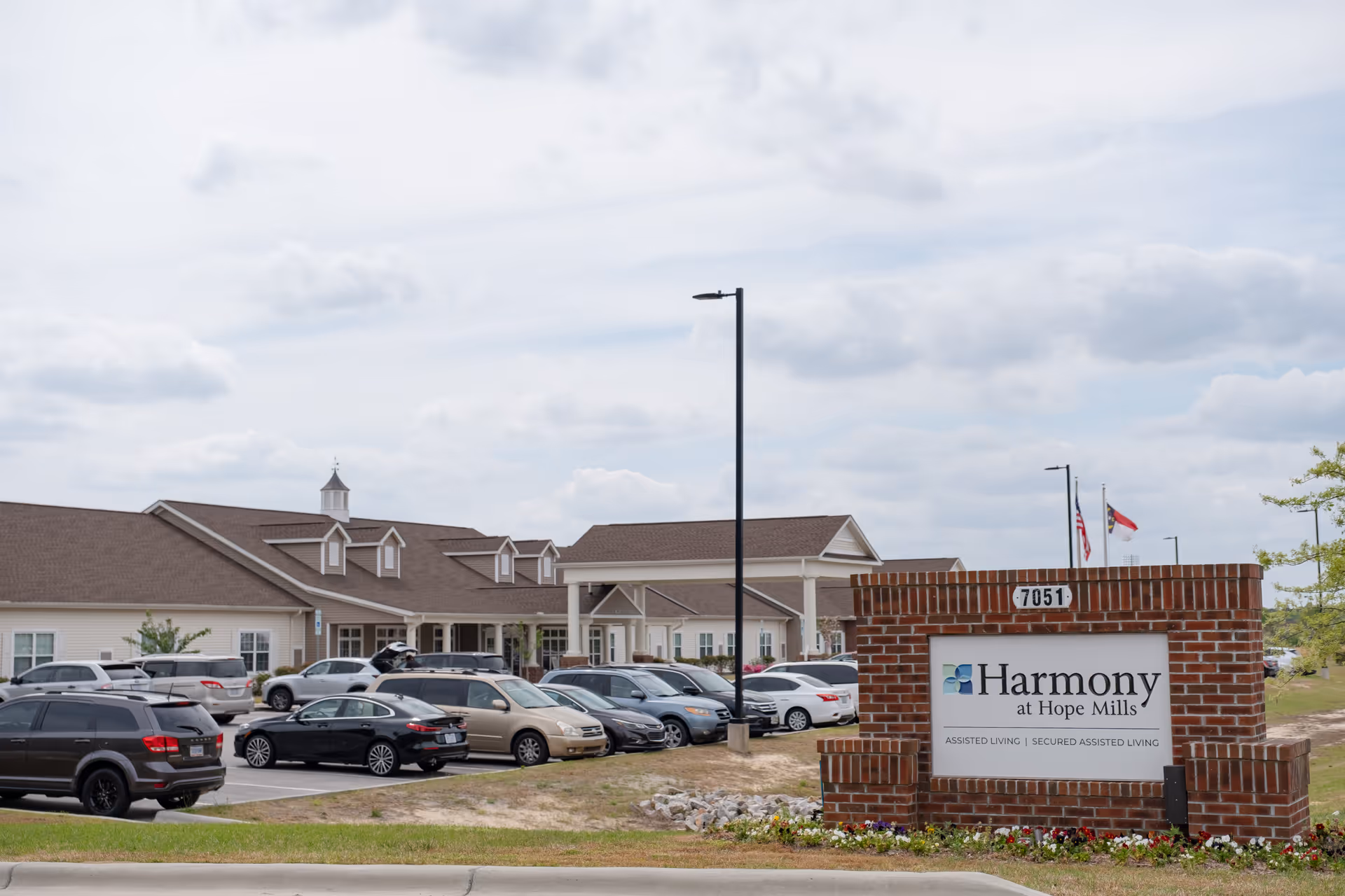 Exterior view of Harmony at Hope Mills assisted living facility showing a brick sign with the facility name and several parked cars in front of a single-story building under a cloudy sky.