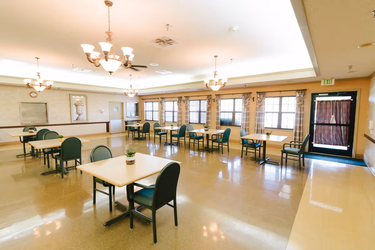 A bright and spacious dining room with multiple square tables and green cushioned chairs arranged neatly. The room has large windows with plaid curtains allowing natural light to enter. There are several ceiling light fixtures and a door with an exit sign above it.
