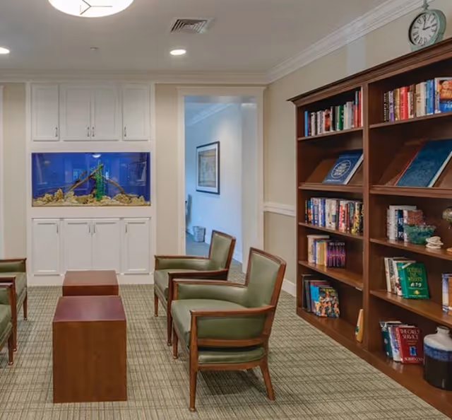 A senior living common room with green upholstered chairs, wooden tables, a bookcase, and a built-in aquarium in a bright interior.