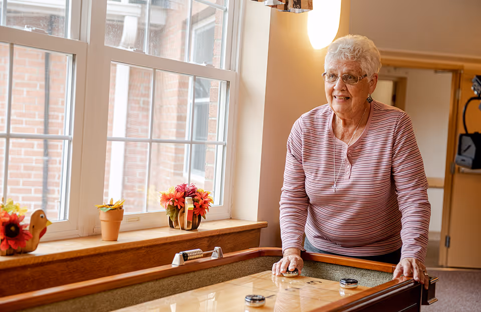 An elderly woman with short white hair and glasses, wearing a striped long-sleeve shirt, is playing shuffleboard indoors near a window with flower decorations on the windowsill.