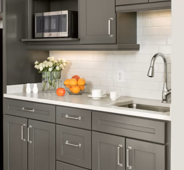 Modern kitchen countertop with gray cabinets, stainless sink and faucet, microwave above, and a bowl of oranges next to a vase of white flowers.