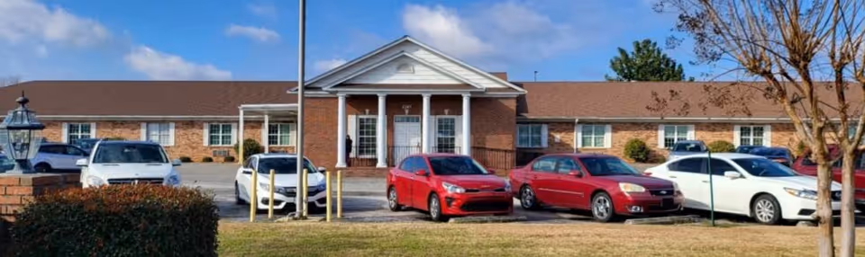 Front exterior view of Honorage Nursing Center, a single-story brick building with a central entrance featuring white columns and a triangular pediment. Several cars are parked in front of the building, and there is a grassy area with a tree and bushes in the foreground under a partly cloudy sky.