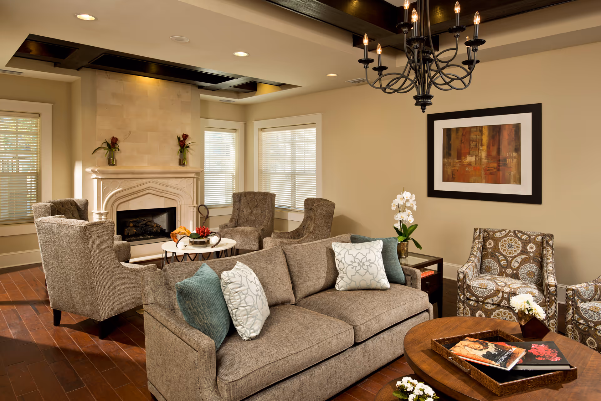 A cozy living room with a beige sofa adorned with decorative pillows, two patterned armchairs, and two gray armchairs arranged around a fireplace. The room features wooden flooring, a round wooden coffee table with books and flowers, a chandelier hanging from the ceiling, and a framed abstract painting on the wall.