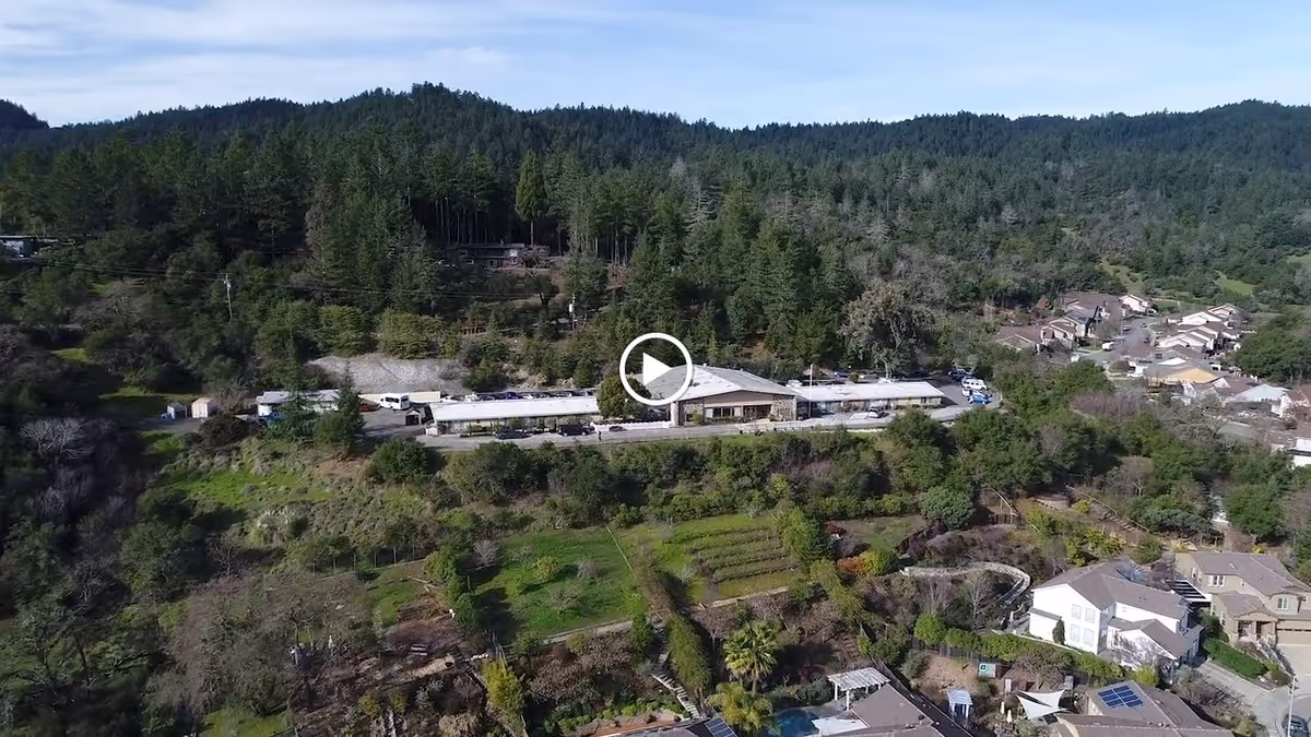 Aerial view of Cloverdale Healthcare Center surrounded by dense forested hills and residential houses, with a clear sky above.