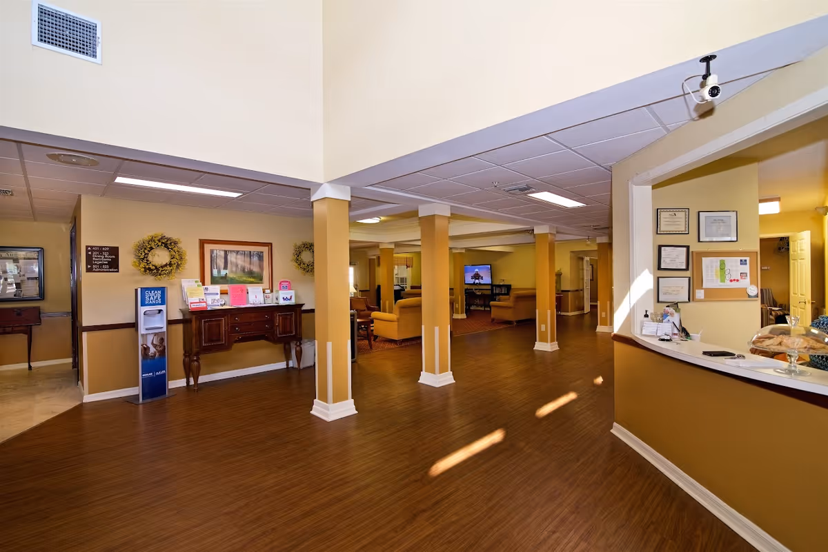 Interior view of a senior living facility lobby area with wooden flooring, beige walls, and several support columns. There is a reception desk on the right with certificates and a bulletin board on the wall behind it. In the background, there are seating areas with armchairs and a television. A hand sanitizer station and informational materials are visible on a wooden console table against the left wall.
