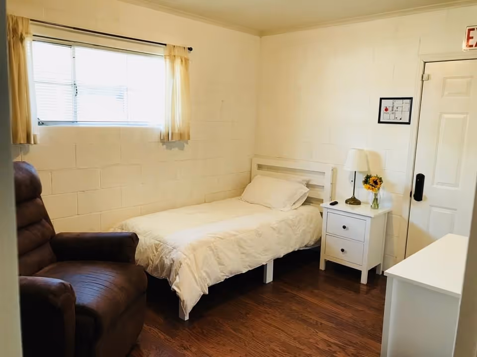 A small, simple bedroom with a single bed covered in white bedding, a brown recliner chair, a white nightstand with a lamp and a vase of sunflowers, a window with beige curtains, and a door with an exit sign above it.