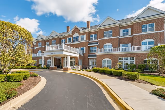 Exterior view of a large three-story brick building with white trim and multiple windows. The building has a covered entrance with columns and a curved driveway leading up to it. Well-maintained landscaping with bushes and trees surrounds the building under a partly cloudy sky.