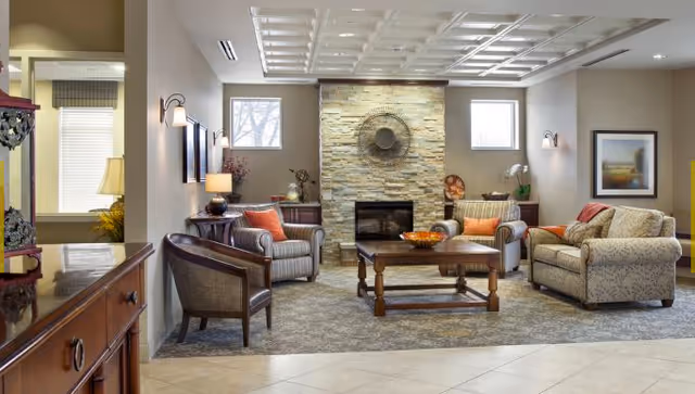 A cozy living room with a stone fireplace centered on the far wall, flanked by two windows. The room features a wooden coffee table with a bowl of fruit, two striped armchairs with orange cushions, a patterned sofa with orange cushions, and a dark wooden chair. The walls are decorated with framed artwork and wall sconces, and the floor has a combination of carpet and tile.
