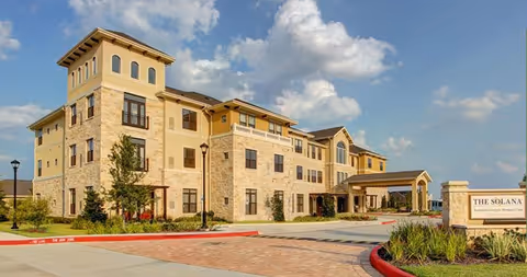 Exterior view of The Solana Vintage Park, a multi-story senior living facility with stone and beige siding, large windows, and a covered entrance. The building is surrounded by landscaped greenery and a paved driveway under a partly cloudy sky.