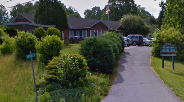 Driveway lined with shrubs leading to a single-story brick assisted living building with parked cars and an American flag in front.