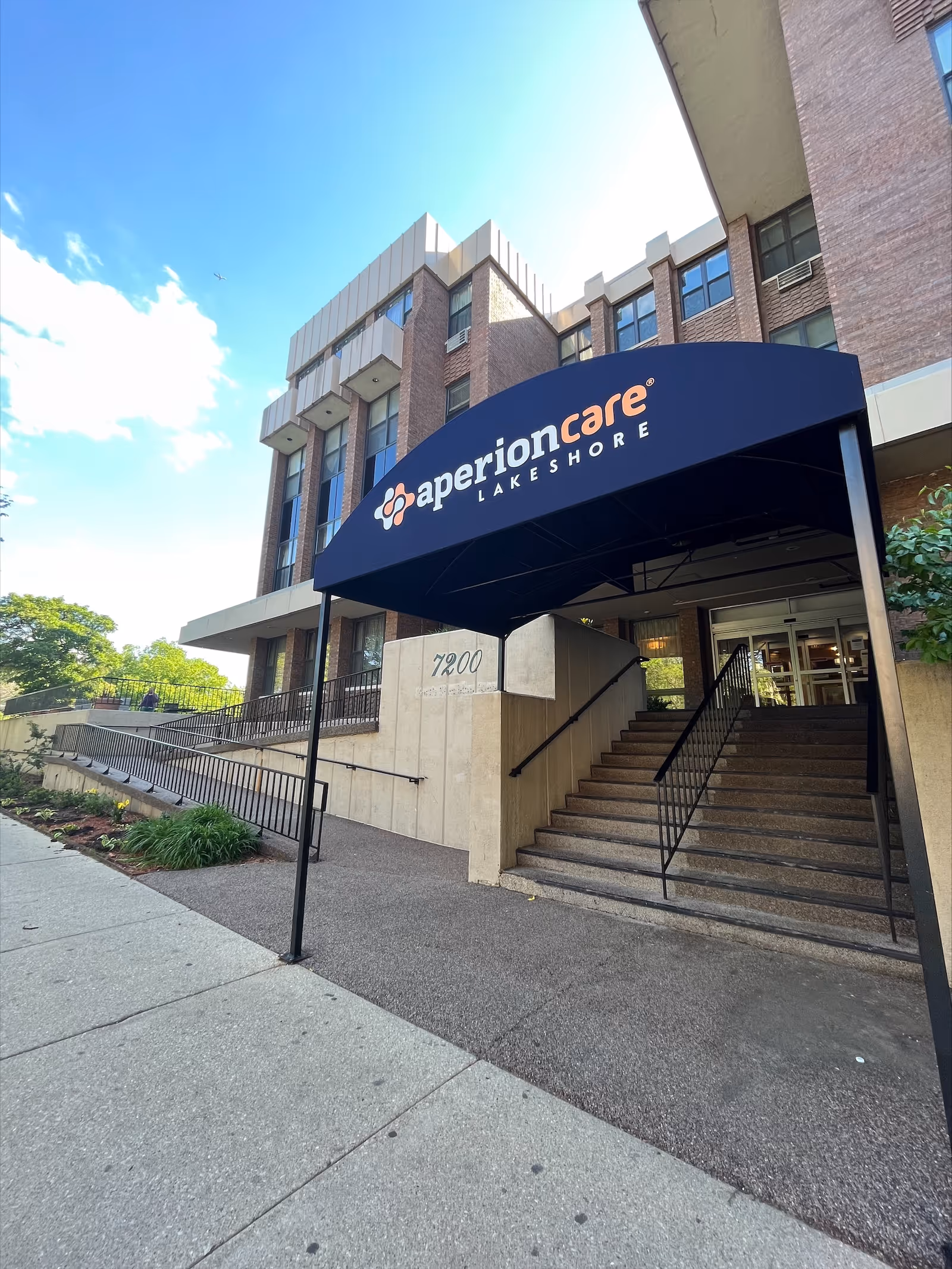 Exterior front entrance of Aperion Care Lakeshore facility showing a brick building with a blue awning displaying the facility name. There are stairs and a wheelchair ramp leading up to the entrance, with some greenery on the side and a clear blue sky above.
