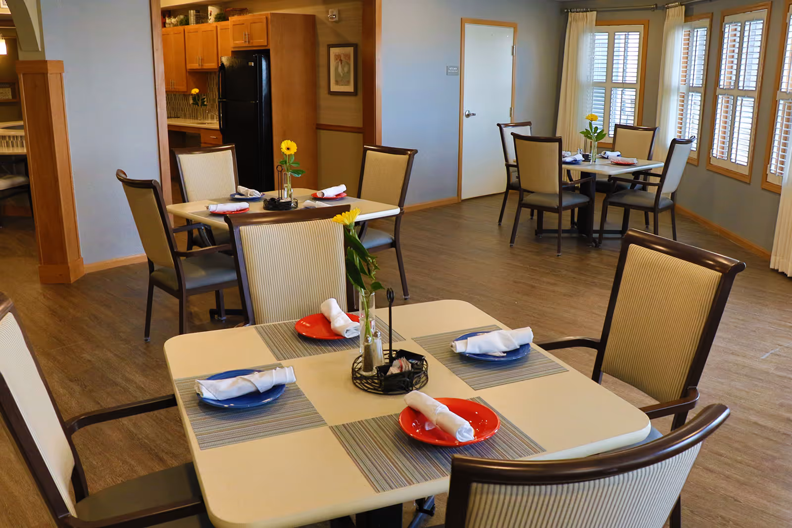 A dining room in a senior living facility with several tables set with plates and rolled napkins. Each table has a small vase with a yellow flower. The room has wooden flooring, light-colored walls, large windows with wooden blinds, and a kitchen area visible in the background.