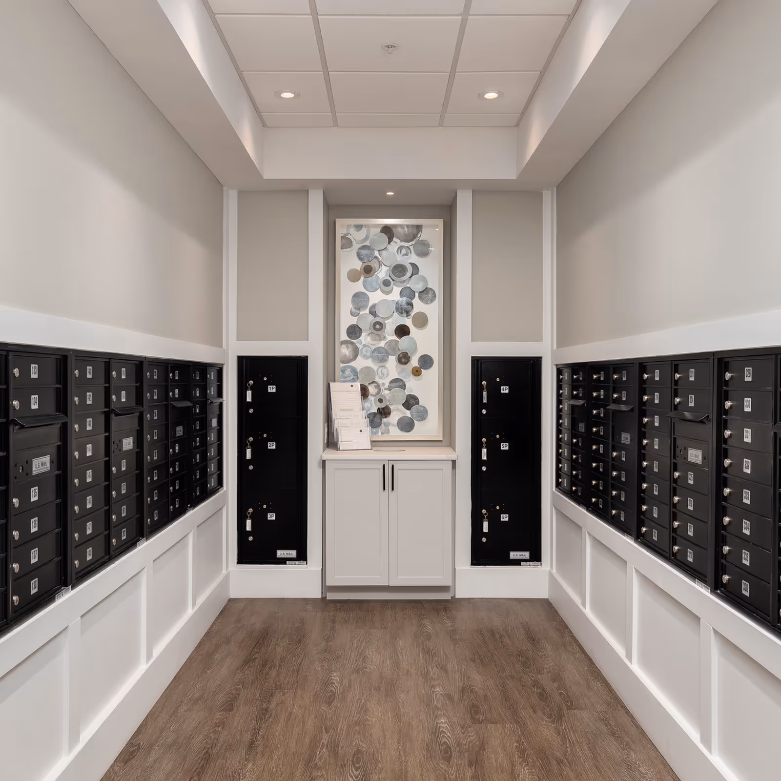 A clean mailroom with rows of black mailboxes lining both walls and a small cabinet with a decorative glass panel at the far end.