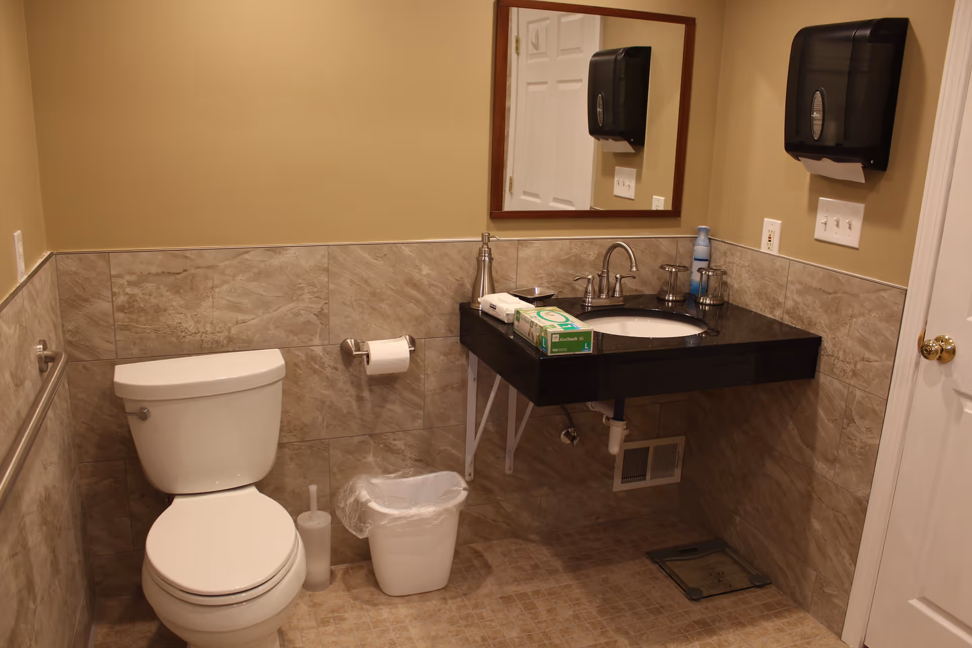 Accessible bathroom with a toilet, wall-mounted sink and mirror, paper towel dispenser, and a small trash can.