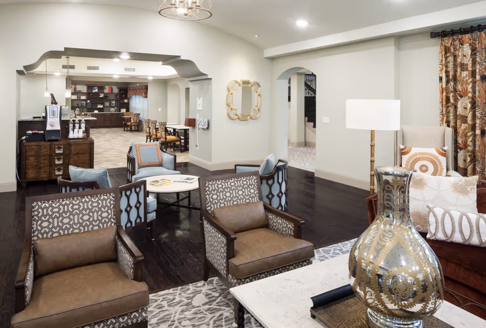 A spacious and well-lit common area in a senior living facility featuring multiple seating arrangements with patterned and leather chairs, a coffee table with a decorative vase, a floor lamp, and floral curtains. In the background, there is a dining area with tables and chairs, and a beverage station with a coffee machine and cups on a wooden cabinet.