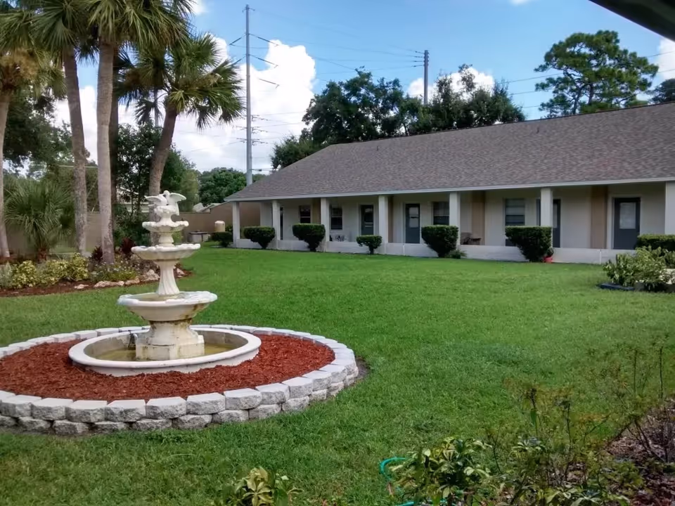 Outdoor view of a senior living facility with a well-maintained lawn, a three-tiered white fountain surrounded by a circular stone border and red mulch, several palm trees, and a single-story building with multiple doors and windows. The sky is partly cloudy.