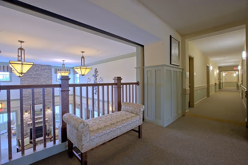 Interior hallway of a senior living facility with beige walls and carpeted floor. A cushioned bench with floral upholstery is placed near a wooden railing overlooking a lower level with a stone fireplace and hanging pendant lights. The hallway extends to the right with several doors and wall-mounted lights.