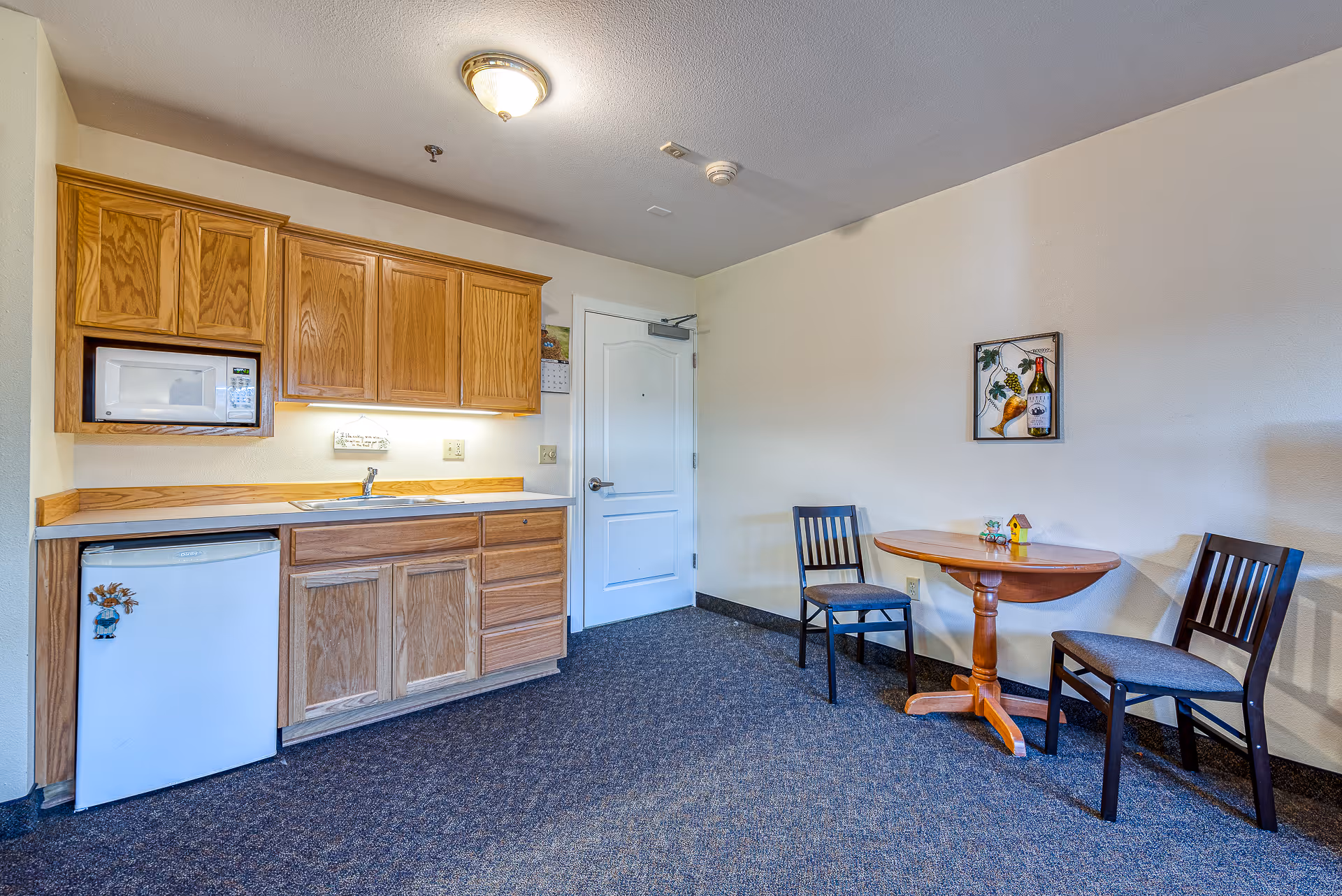 Small assisted-living kitchenette with wooden cabinets, a sink, microwave, mini-fridge and a round table with two chairs.