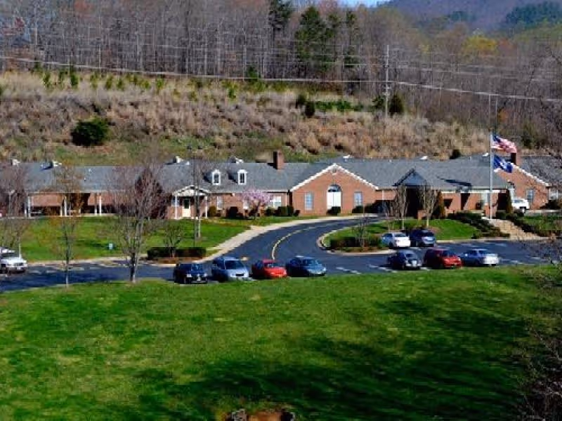 A single-story brick building with a gray roof, surrounded by a parking lot with several cars parked. There is a large green lawn in the foreground and a wooded hillside in the background. Two flagpoles with flags are visible near the building entrance.