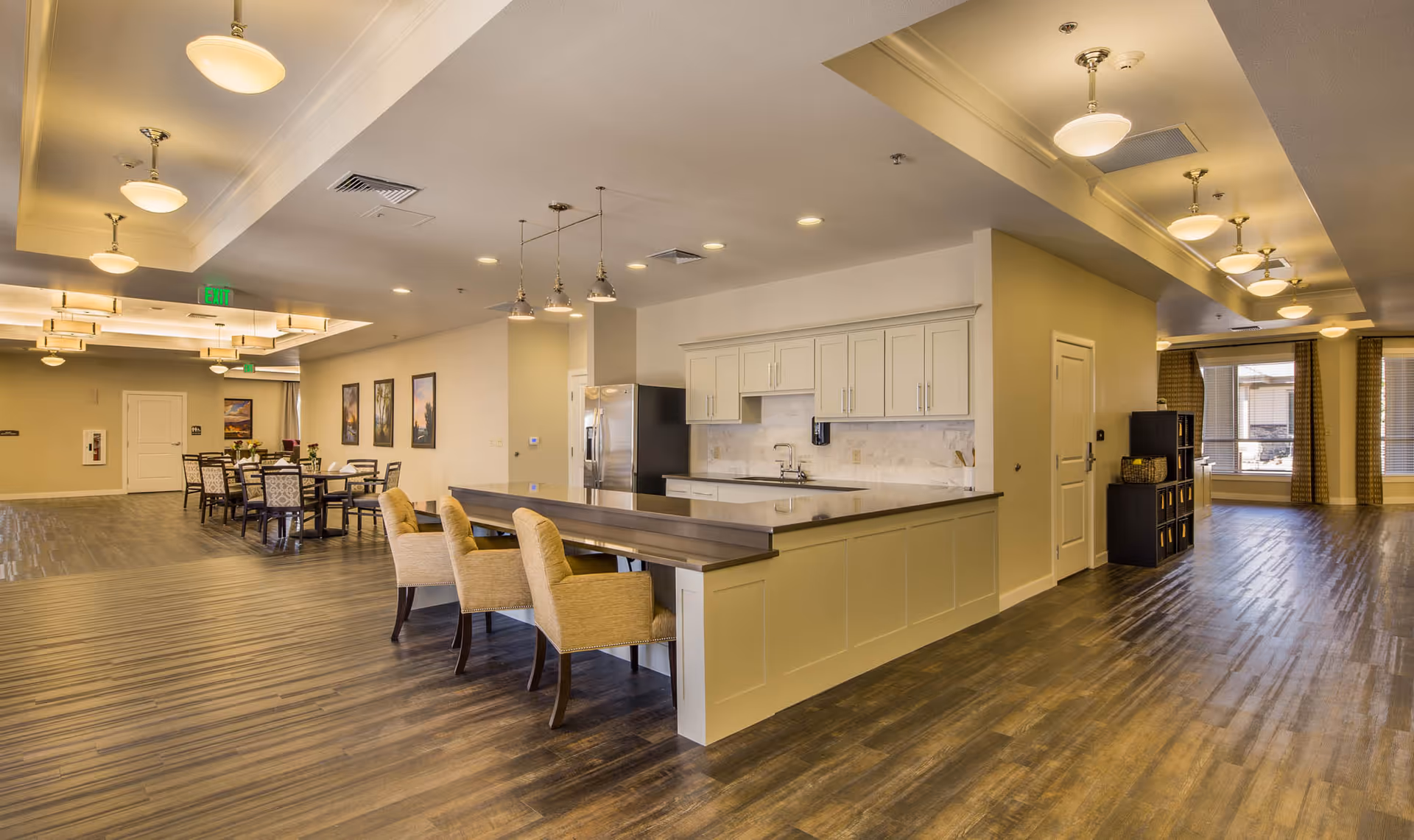 Spacious interior of a senior living facility featuring a modern kitchen area with white cabinets, a stainless steel refrigerator, and a long counter with beige upholstered chairs. Adjacent to the kitchen is a dining area with multiple tables and chairs, warm lighting fixtures on the ceiling, and framed artwork on the walls. Large windows with curtains allow natural light into the space.