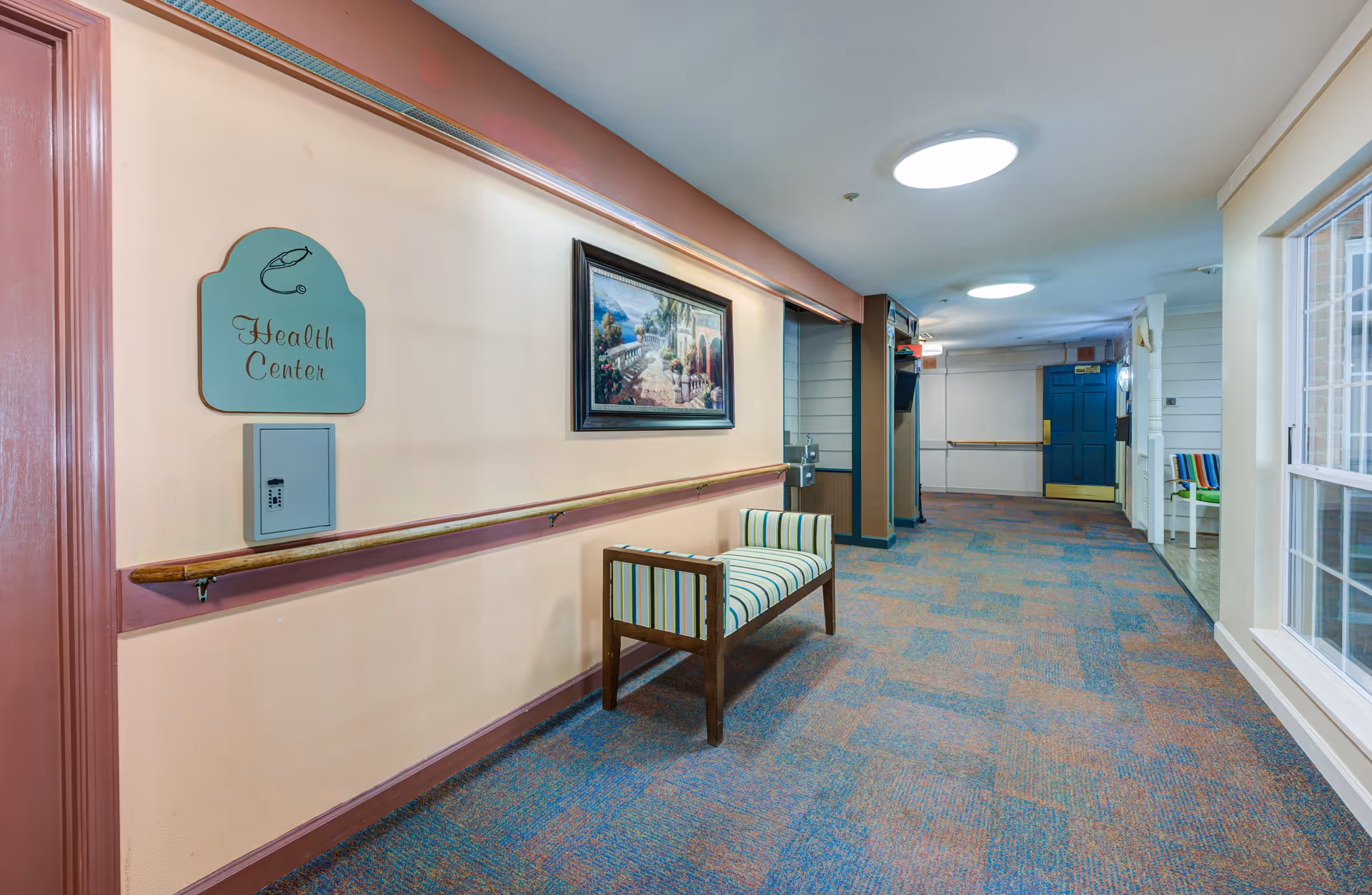 A hallway in a senior living facility with a sign labeled 'Health Center' on the left wall. The hallway has a striped bench, a framed painting, handrails along the walls, and a carpeted floor. There are large circular ceiling lights and a window on the right side letting in natural light.