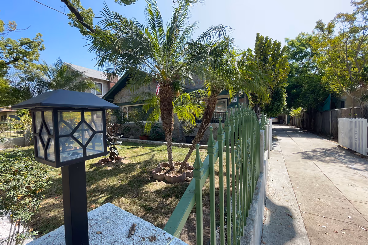 Exterior view of a residential-style building with a stone facade partially obscured by palm trees and a green metal fence. A black lantern-style light fixture is mounted on a white stone post in the foreground. The scene is sunny with a clear blue sky and a sidewalk running alongside the property.