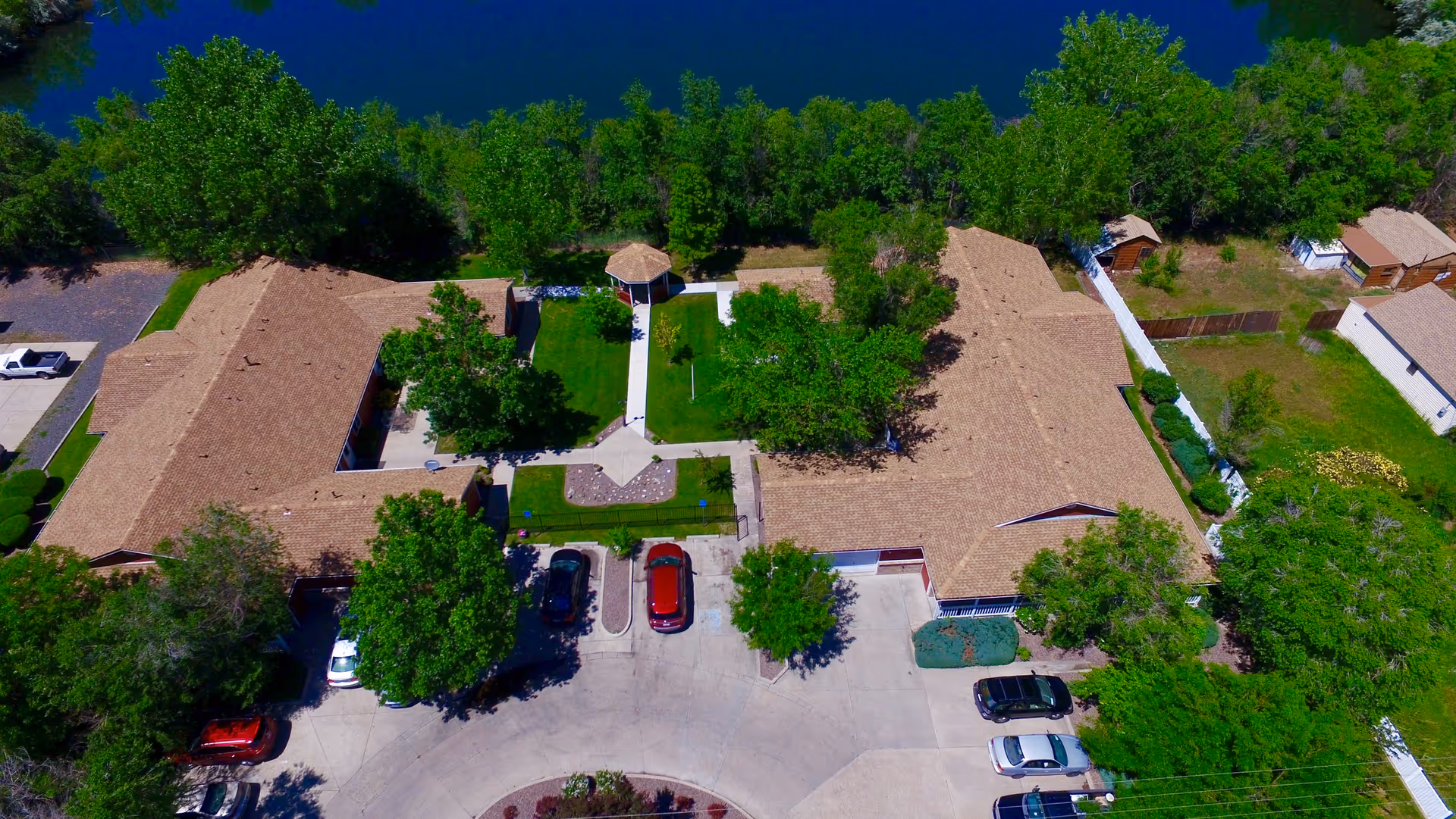 Aerial view of The Verandas Assisted Living facility showing two large buildings with brown roofs surrounding a green courtyard with trees and a gazebo. Several cars are parked in the driveway and parking areas around the buildings. The facility is bordered by trees and a body of water is visible at the top of the image.