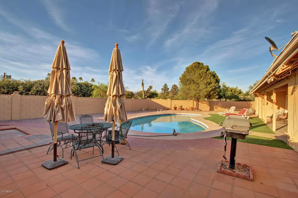 Backyard patio with a table and closed umbrellas, a grill, and a swimming pool with lounge chairs and a fence.