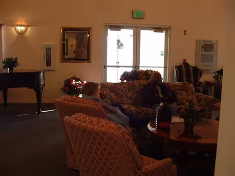 A cozy living room area in a senior living facility with two elderly people seated on patterned sofas around a wooden coffee table. There is a grand piano on the left side, framed artwork on the wall, and double glass doors with an exit sign above them in the background.