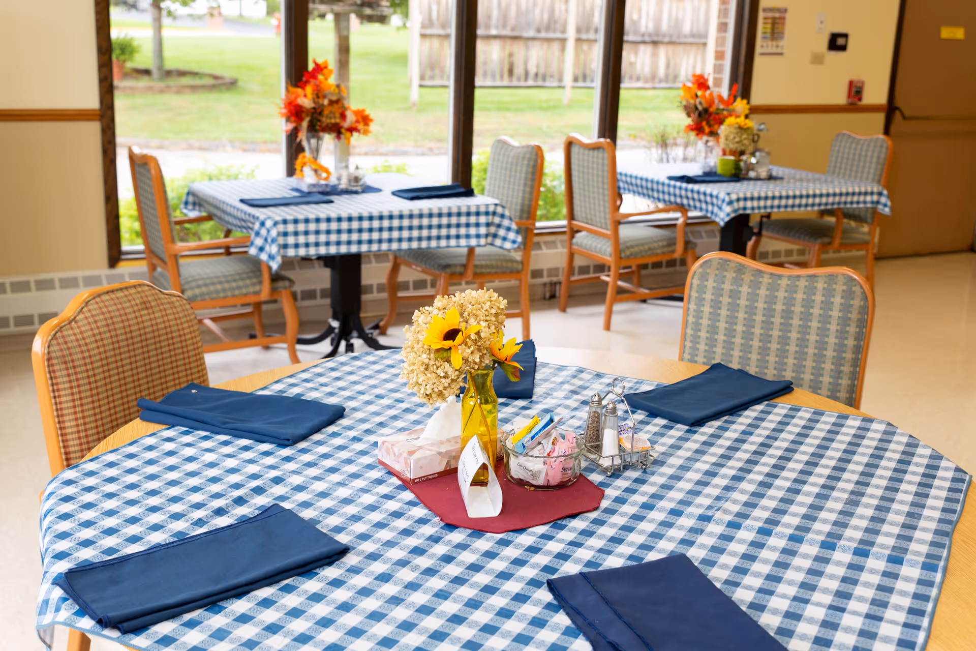 Dining room with round tables covered in blue gingham tablecloths, napkins, and floral centerpieces by large windows.