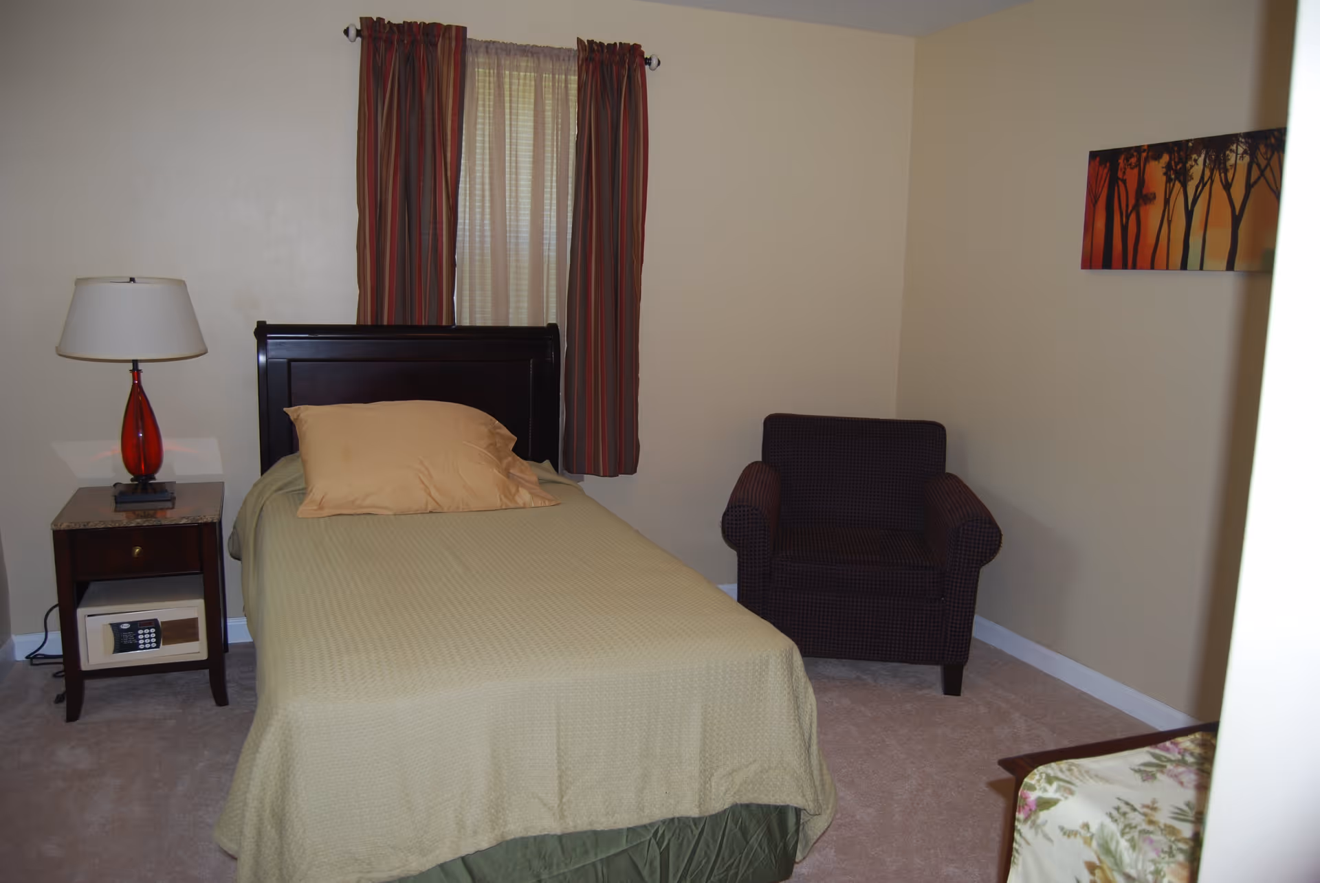 A small bedroom with a single bed covered in a beige bedspread and a tan pillow. Next to the bed is a wooden nightstand with a red lamp and a small safe underneath. A window with striped curtains is behind the bed. To the right of the bed is a dark upholstered armchair, and a framed artwork of trees with a sunset background hangs on the wall.
