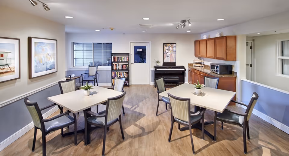 Communal dining/activity room with multiple tables and chairs, a kitchenette area, bookshelf and piano.
