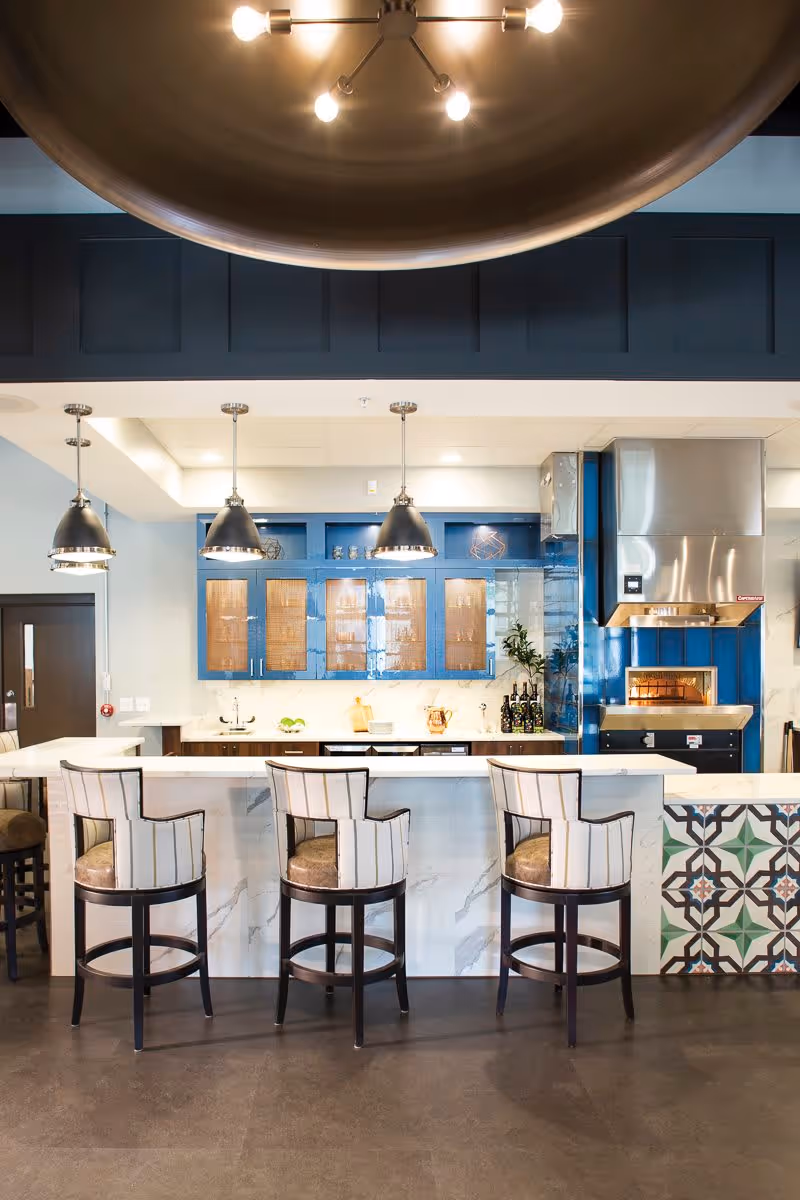 Modern kitchen area with a white marble island countertop and three high-back chairs with striped upholstery. Above the island, there are three pendant lights. The back wall features blue cabinets with glass doors, a stainless steel oven hood, and a built-in oven. The right side of the island has decorative tile accents.