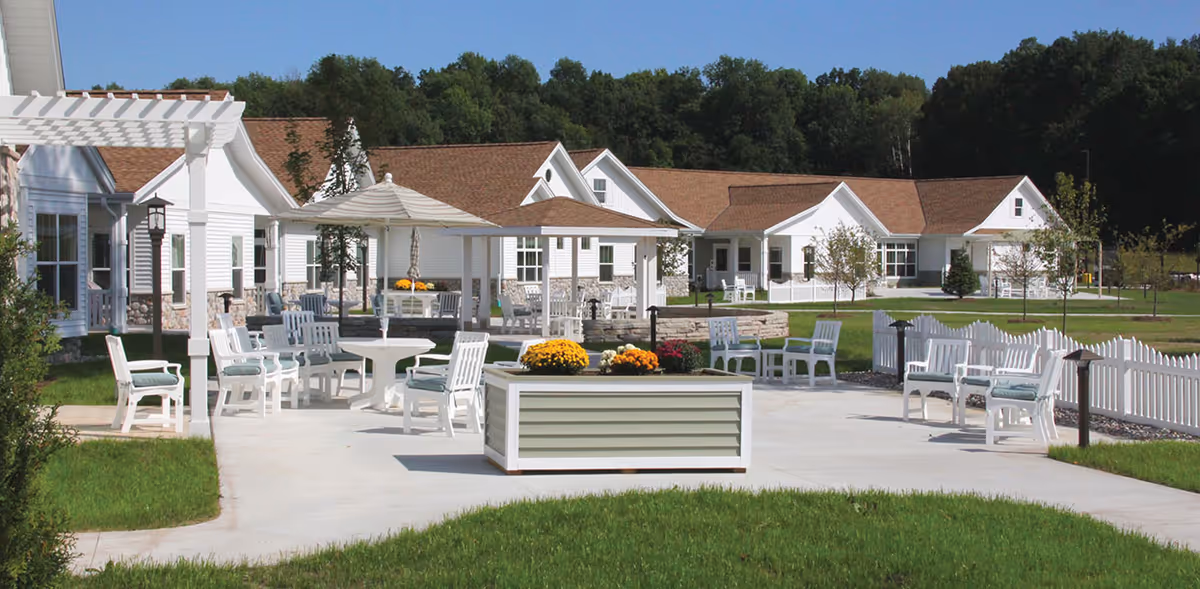 Outdoor courtyard with white chairs, tables, a pergola and planter boxes in front of single-story cottage buildings